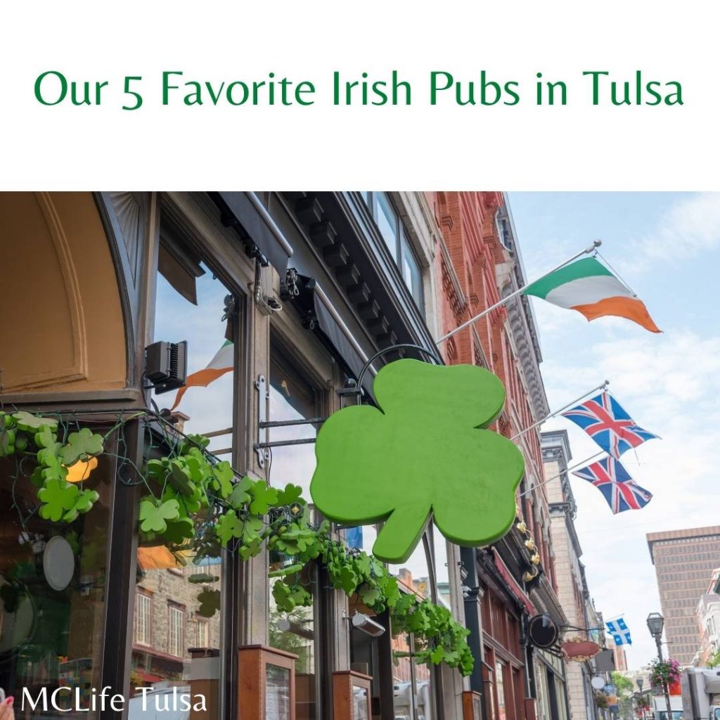 Image of a green clover sign outside of a bar.  In the background there are colorful flags outside of the bar, and a party cloudy sky in the background.  